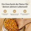 Nahaufnahme eines Glases mit goldgelbem Bienenbrot (Perga), in das eine Holzschaufel eingetaucht ist. Darüber ein Banner mit der Aufschrift 